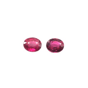 May include: Two oval-cut, deep red gemstones with a polished surface. The gems are set against a white background, showcasing their vibrant color and clarity. These gemstones are likely rubies, often used in jewelry.