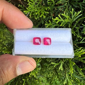 May include: Two vibrant, cushion-cut red gemstones are displayed side-by-side within a clear, rectangular plastic storage case. The case features a white, soft-textured interior insert with two parallel cylindrical slots to hold the stones securely.