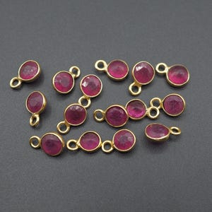 May include: Gold-toned jewelry findings with faceted, round, magenta-colored gemstones. The findings include single pendants and linked sets of two and three gemstones. The jewelry components are displayed on a dark gray surface.