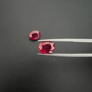 May include: Two oval-cut, deep red rubies with faceted surfaces. One ruby is held by a pair of silver-toned metal tweezers, while the other ruby rests nearby. Both gemstones exhibit a vibrant, saturated crimson color and a polished finish.