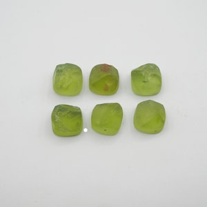 May include: Six green peridot gemstones, cut into square shapes, are arranged in two rows of three on a white surface.