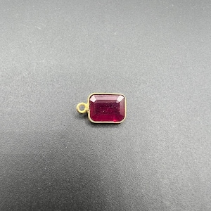 Puede incluir: Un colgante rectangular con una piedra preciosa facetada de color rojo intenso engastada en un marco de metal dorado. El colgante incluye una pequeña anilla circular dorada unida a un lado, diseñada para conectarse a una cadena o cordón.