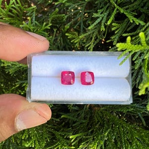 May include: Two cushion-cut, vibrant pinkish-red gemstones are displayed side-by-side in a clear plastic storage box with a white foam insert. The faceted stones exhibit a square shape with rounded corners and a polished, reflective surface.