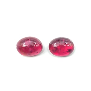 May include: Two polished, oval-shaped, deep red gemstones. The stones have a smooth, glossy surface and a translucent appearance, with subtle internal inclusions. The gems are set against a bright white background.