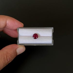 May include: A round, deep red ruby gemstone displayed in a small, clear plastic case with a white interior. The gemstone is the focal point, set against a dark background. The case is held by a hand.