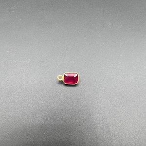 以下が含まれることがあります： ゴールドトーンの金属フレームにセットされた深い赤色の宝石が特徴の小さな長方形のペンダント。宝石は光を反射するファセット加工された表面を持ち、吊り下げるための小さな円形のゴールドトーンの丸カンが片側に付いています。