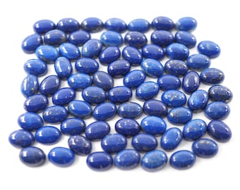 Puede incluir: Una colección de piedras preciosas de lapislázuli ovaladas y pulidas. Las piedras son de un color azul intenso y rico con motas doradas, y están dispersas sobre una superficie blanca. Estas piedras son ideales para la fabricación de joyas.