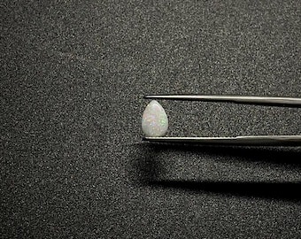 May include: A pear-shaped, iridescent opal gemstone is held by a pair of tweezers against a black background. The opal displays a milky white base color with flashes of green, blue, and pink. The tweezers are silver-colored.