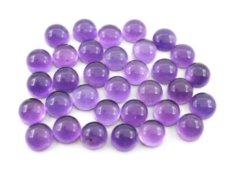 Puede incluir: Colección de piedras preciosas redondas y pulidas de amatista. Cada gema es de color púrpura intenso con una superficie lisa y reflectante. Las piedras están dispuestas juntas sobre un fondo blanco.