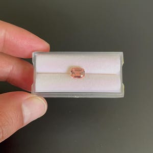 Puede incluir: Una gema de color melocotón, de forma rectangular, se muestra dentro de una caja de plástico transparente con un interior blanco. La gema está sujeta por los dedos de una persona sobre un fondo oscuro.