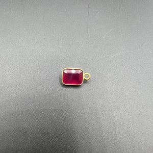 以下が含まれることがあります： 角が丸いゴールドトーンの金属フレームに、深い赤色の宝石がセットされた長方形のペンダント。ペンダントの右側には、チェーンやジュエリーパーツに取り付けるための小さな円形の丸カンが付いています。