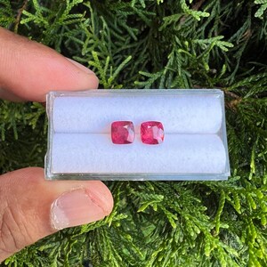 May include: Two loose, cushion-cut, vibrant pink gemstones displayed side-by-side in a clear, rectangular plastic storage box with a white, padded interior insert. The stones feature faceted surfaces that reflect light, showcasing a deep, saturated color.