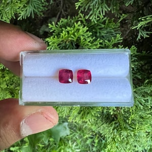 May include: Two cushion-cut, vibrant pinkish-red gemstones are displayed side-by-side within a clear, rectangular plastic storage case. The case features a white, soft-textured foam insert with two horizontal slots designed to hold the loose stones securely.