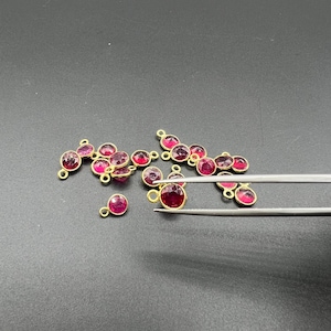 May include: A collection of small, round, faceted dark pink gemstones set in gold-toned metal bezels, each featuring a tiny circular loop at the top for attachment. One of these individual jewelry charms is being held by silver-toned metal tweezers.