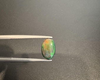 May include: An oval-shaped cabochon opal gemstone displaying a dark base color with vibrant flashes of green, orange, and yellow play-of-color. The gemstone is held by a pair of silver-toned metal tweezers, showcasing its polished, smooth surface.