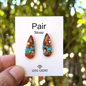 May include: A pair of teardrop-shaped stone earrings. The stones feature a mix of colors, including turquoise, orange, and brown. The earrings are displayed on a white card with the words "Pair Stone" and the "GTG GEMS" logo.