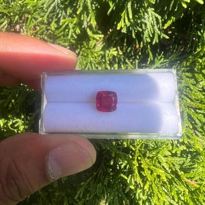 May include: A cushion-cut ruby gemstone with a deep red color, securely placed in the center of a small, rectangular, clear plastic display box featuring a white velvet-like insert with two horizontal slots for holding the loose stone.