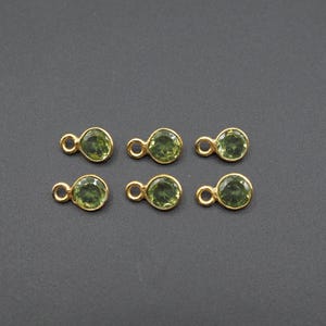 May include: Six small, round, gold-toned jewelry charms, each featuring a faceted, olive-green gemstone set in a bezel. Every charm includes a small, circular jump ring attached to the side for easy connection to chains or other jewelry components.