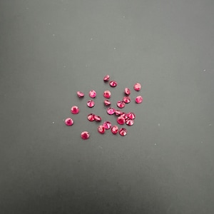 May include: A collection of small, round, faceted loose gemstones in a vibrant, deep pinkish-red hue. These individual stones are scattered across the frame, each displaying multiple facets that catch the light to create a sparkling effect.