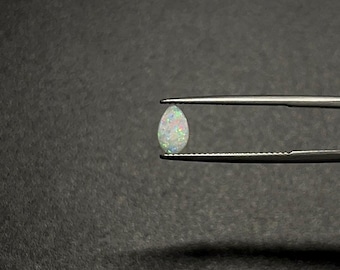 May include: A pear-shaped opal gemstone held by tweezers. The opal displays a milky white base color with flashes of iridescent pink, blue, and green. The gemstone is set against a dark background.