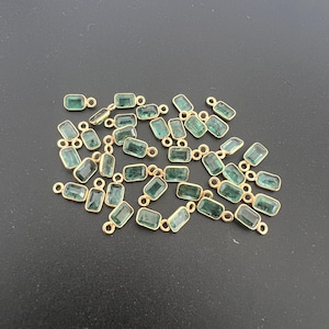 May include: A collection of rectangular emerald green gemstone charms with gold-colored metal settings. Each charm has a small loop at the top for attaching to jewelry. The charms are scattered on a dark surface.
