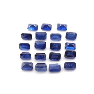 May include: A collection of twenty rectangular, deep blue gemstones, possibly sapphires, arranged on a white surface. Each gem has a polished, faceted appearance, reflecting light. The gems are of similar size and shape.