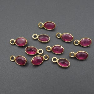 May include: A collection of gold-colored jewelry findings, each featuring an oval-shaped, deep red gemstone. The findings have a small loop at the top for attaching to other jewelry components. The gemstones are set in a gold bezel.