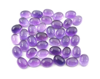 Puede incluir: Una colección de piedras preciosas de amatista ovaladas en tonos de púrpura, desde lavanda claro hasta violeta intenso. Las piedras pulidas están dispersas sobre una superficie blanca, mostrando su textura suave y brillante. Ideal para joyería o coleccionismo.