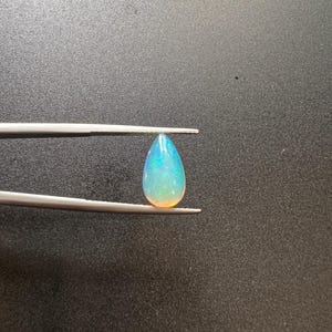 May include: A pear-shaped opal gemstone displaying iridescent flashes of blue, green, and orange light. The translucent stone is held by a pair of silver-toned metal tweezers, showcasing its smooth, polished surface and vibrant play of color.