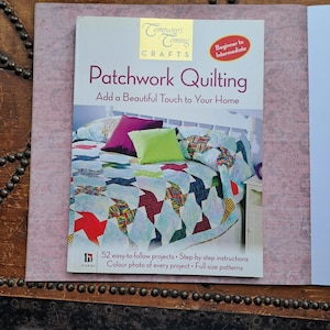 May include: A softcover instructional book titled Company's Coming Crafts Patchwork Quilting. The cover features a bed with a colourful patchwork quilt, a purple pillow, and a lime green pillow. Text includes 52 easy-to-follow projects and patterns.