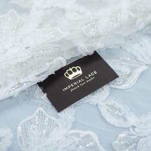 Puede incluir: Primer plano de tela de encaje floral blanco con detalles intrincados y acentos brillantes. Una tarjeta de visita negra con texto dorado que dice "IMPERIAL LACE french lace dealer" se coloca sobre la tela. El encaje está sobre una superficie azul claro.