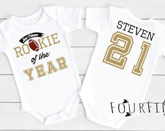 Puede incluir: Dos bodies blancos para bebé. Uno presenta un balón de fútbol y el texto "New Orleans Rookie of the Year" en dorado y negro. El otro tiene el nombre "STEVEN" y el número "21" en dorado y negro. La marca "FOURFINS" está impresa en la parte inferior.