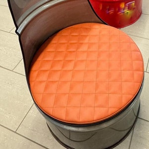Könnte beinhalten: Ein grauer Metallstuhl mit einem orangefarbenen, gesteppten Sitzkissen. Der Stuhl ist aus einem wiederverwendeten Metallfass gefertigt.
