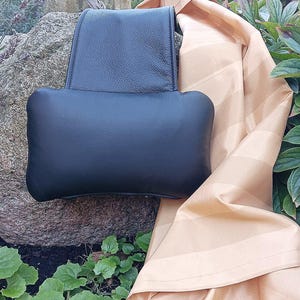 Könnte beinhalten: Ein schwarzes Leder-Kopfstützenkissen mit rechteckiger Form. Das Kissen liegt auf einem Stein mit grünen Pflanzen im Hintergrund.