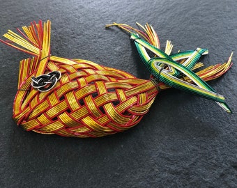 May include: A gold, red, and green woven fish ornament with a black knot on the body. The fish is made of braided cord and has a long tail.