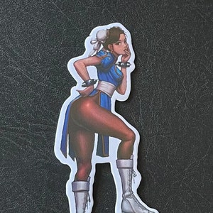 May include: A sticker featuring a female character with brown hair, wearing a blue and white outfit, white boots, and a white headband. The character is posed with one hand on her hip and the other hand on her chin.