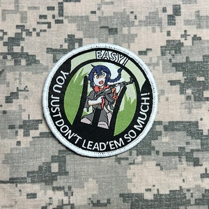 May include: A round, white-bordered patch with a black background. The patch features a cartoon girl with blue hair holding a rifle. The text on the patch reads "You Just Don't Lead 'Em So Much! Easy!"