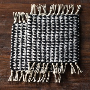 May include: Two black and white woven coasters with fringe. The coasters have a geometric pattern and are made of a natural fiber.
