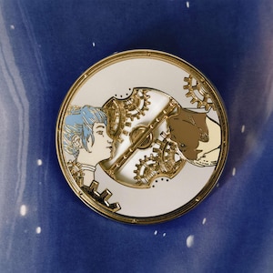 May include: Gold-toned enamel pin featuring a white background with a detailed illustration of a woman and a wolf's head, both partially obscured by a set of interlocking gears.