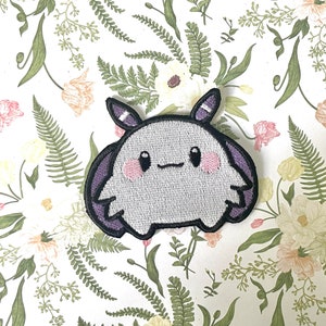 May include: A purple and white embroidered patch of a cartoon creature with large eyes and a smiling mouth. The creature has a rounded body and two small arms.
