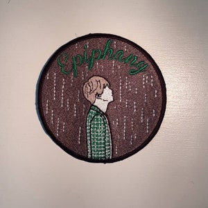 May include: A brown embroidered patch with the text "Epiphany" in green lettering. The patch features a cartoon illustration of a person with brown hair wearing a green and white plaid shirt standing in front of a brown background with white lines.