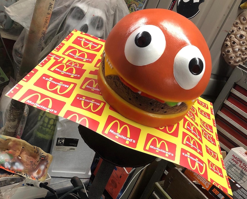 May include: A large, orange, cartoon-style hamburger with googly eyes and a wide, toothy grin. The hamburger is sitting on a yellow and red surface with the text "Morfhead's" repeated.