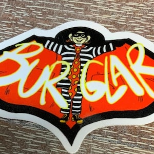 May include: A red and black sticker with the word "BURGLAR" written in white letters. A cartoon character wearing a black and white striped shirt and a red tie with white polka dots is in the center of the sticker.