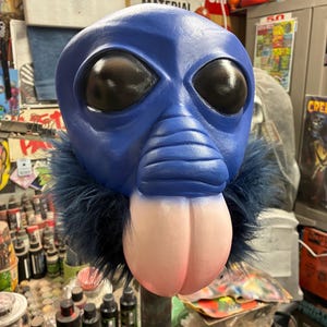 May include: A blue alien mask with large black eyes and a pink, bulbous lower face. The mask has a dark blue fur collar. The mask is mounted on a metal stand, with various paint bottles in the background.