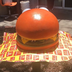 May include: A large, orange, plastic hamburger replica with a yellow cheese slice and a brown patty. The hamburger is sitting on a yellow and red tablecloth with the McDonald's logo repeated.