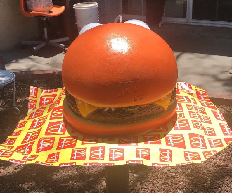 May include: A large, orange, plastic hamburger replica with a yellow cheese slice and a brown patty. The hamburger is sitting on a yellow and red tablecloth with the McDonald's logo repeated.