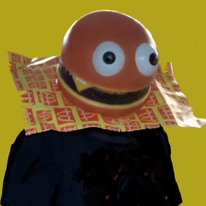 May include: A person wearing a brown and yellow hamburger costume with googly eyes. The costume is made of a soft, squishy material and has a large, toothy grin. The person is wearing a black shirt underneath the costume.