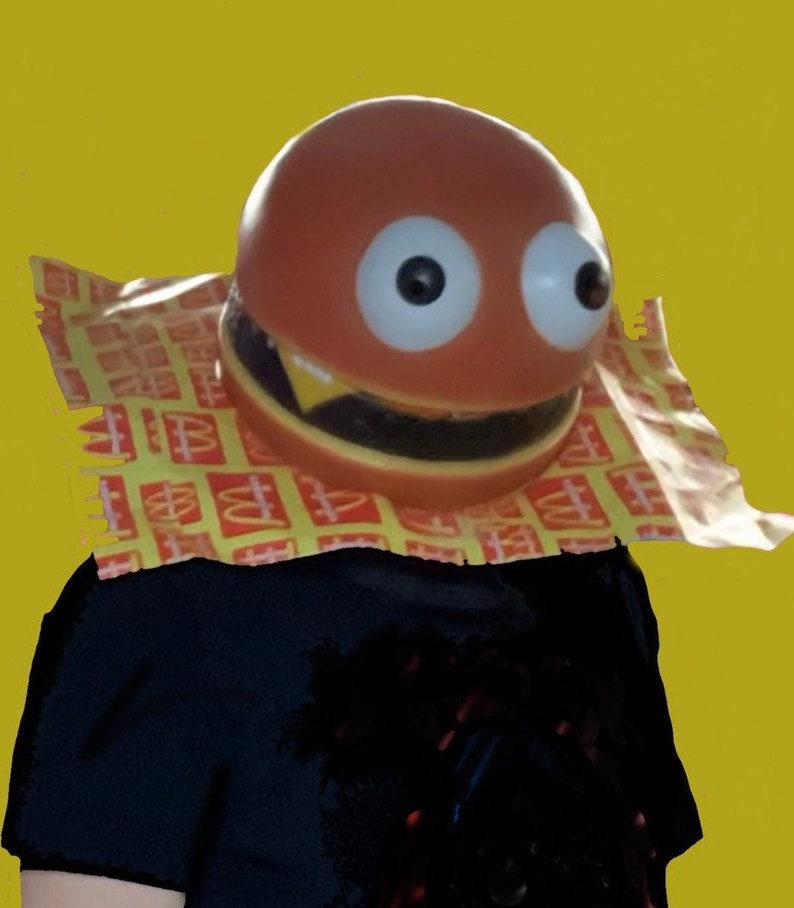 May include: A person wearing a brown and yellow hamburger costume with googly eyes. The costume is made of a soft, squishy material and has a large, toothy grin. The person is wearing a black shirt underneath the costume.