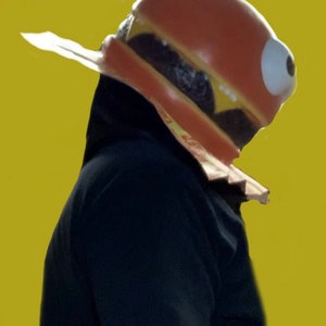 May include: A person wearing a large, red, yellow, and brown hamburger costume with a single large eye. The costume is made of foam and has a realistic appearance.