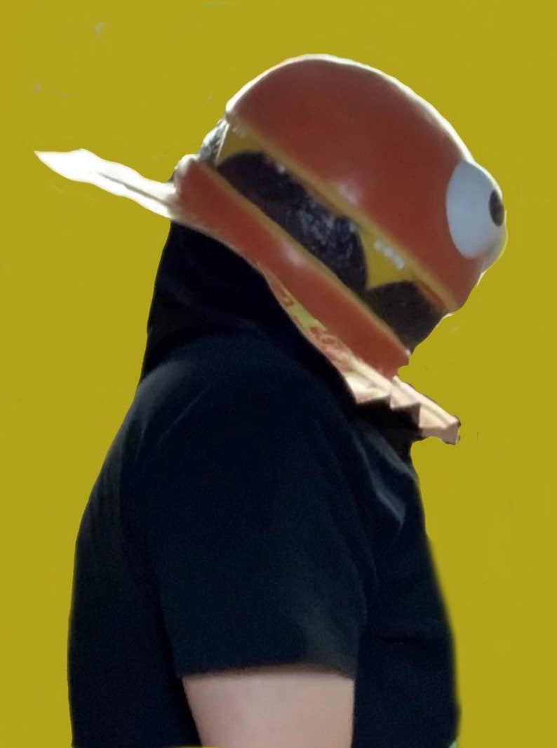 May include: A person wearing a large, red, yellow, and brown hamburger costume with a single large eye. The costume is made of foam and has a realistic appearance.